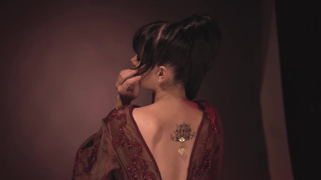 delightful girl with delicate skin and tall horse tails hands her neck, hair and face, showing off a gorgeous tattoo depicting a sweet lotus adorned with a shiny brooch on her open back