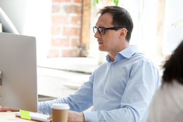 Fototapeta premium Serious businessman, boss in glasses looking at monitor screen squinting, working with document, writing ecenomic report, marketing plan, watching video webinar, typing business email to client