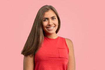 Fototapeta premium A beautiful woman has an attractive white smile isolated on a pink background. A joyful brunette with dark skin in a summer red t-shirt looks at the camera