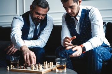 Your move! Two young handsome men in full suits playing chess and smiling while sitting indoors.
