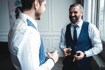 Relaxing. Full length of two young handsome men in suits holding glasses and looking at each other while resting indoors.