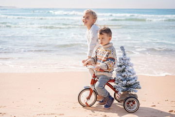 Santa claus siblings kids in Christmas sweater. Happy child on the bicycle or tricycle near water. Xmas party celebration, childhood. Winter holiday vacation. New year small boy at sea beach.