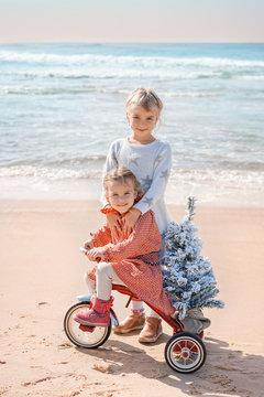 Santa Claus Siblings Kids In Christmas Sweater. Happy Child On The Bicycle Or Tricycle Near Water. Xmas Party Celebration, Childhood. Winter Holiday Vacation. New Year Small Girls At Sea Beach.