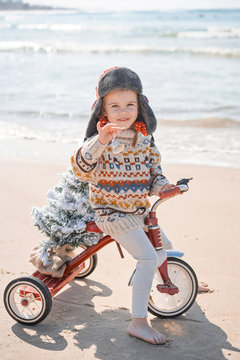 Santa Claus Kid In Christmas Sweater. Happy Child On The Bicycle Or Tricycle Near Water. Xmas Party Celebration, Childhood. Winter Holiday Vacation. New Year Small Girl At Sea Beach.