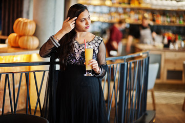 Pretty indian girl in black saree dress posed at restaurant with orange juice at hand.