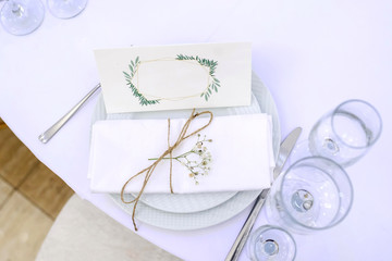 Obraz na płótnie Canvas Closeup photo of a set of two plates: one above the other, near knifes, forks and glasses with a cotton white napkin inwrought in a beige vintage thread with a flower on a white tablecloth