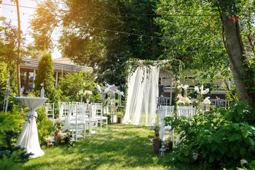 Rows of white empty chairs on a lawn near a wedding round arch decorated with white curtains and...