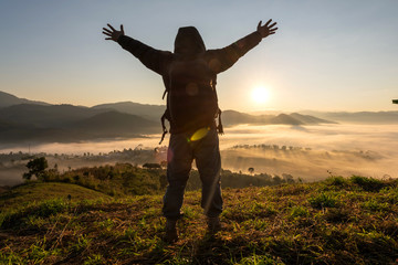 Silhouette of happy tourist man standing on top mountain with raised hands and looks into the distance at morning sunrise with foggy mountain landscape