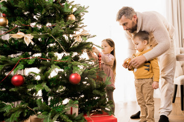 Warm-toned portrait of happy father decorating Christmas tree with two adorable kids, copy space