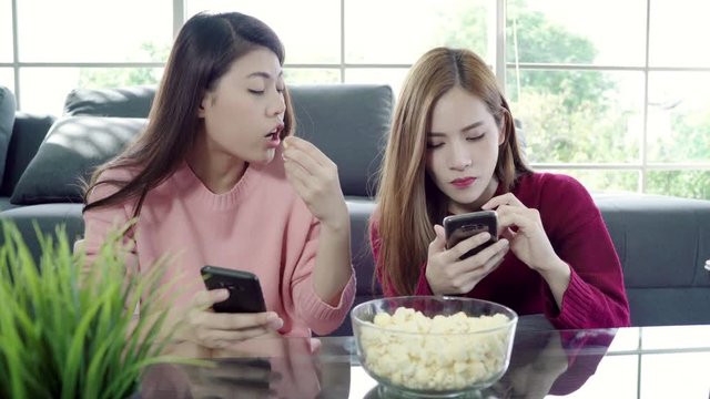 Asian women using smartphone and eating popcorn in living room at home, group of roommate friend enjoy funny moment while lying on the sofa. Lifestyle women relax at home concept.