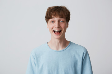 Close up of amazed excited young man with short disheveled hair and braces on teeth wears blue t-shirt shouting and feels happy surprised isolated over white background