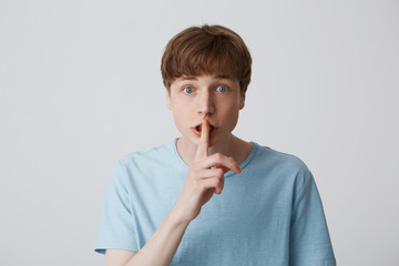 Young guy with wide open eyes, keeps fore finger on lips, calls to keep a secret, do not tell anyone, keep quiet, do not make noise, demonstrates silence gesture, isolated over white background