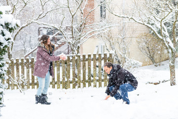 Young man, woman playing, making, sitting, standing, throwing snowballs in winter snowstorm, storm, snowing at home, house garden, front yard, backyard, trees covered in snow, smiling, happy, laughing
