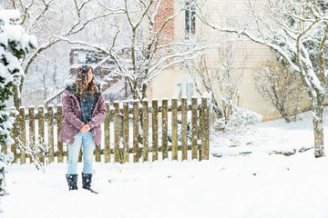 Young woman standing in winter coat in front yard, backyard with covered trees by house, wooden fence at snow, snowing snowstorm, storm, enjoying weather, falling snowflakes