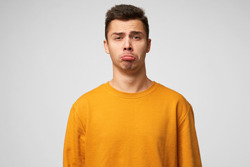 Expresses insult childly. Portrait of emotional guy looks sad, upset, frustrated, displeased, lower lip pouted, showing defenseless, wears orange sweatshirt, stands isolated over white background.