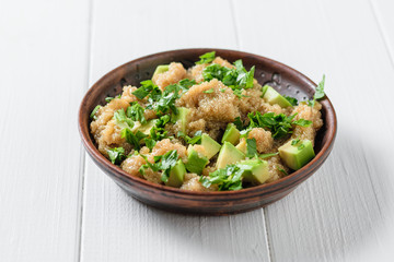 Clay bowl with amaranth seed salad with avocado slices and parsley on white wooden table.
