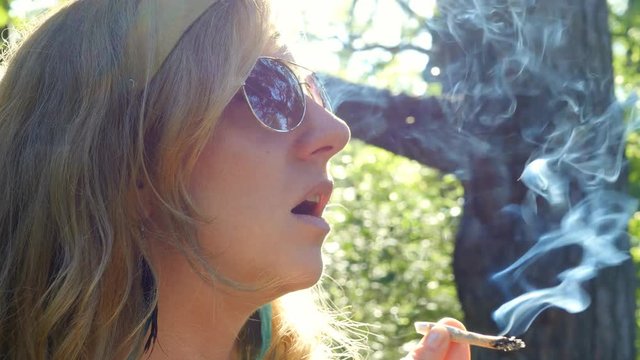Young blonde woman with sunglasses takes drag from marijuana joint. She inhales and then exhales smoke. Profile view.