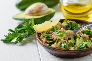 Clay bowl with amaranth seed salad with avocado slices, lemon and parsley on white wooden table.