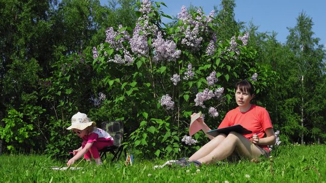 A young mother with dark hair and a cute little daughter paint next to a blooming lilac in the park on a spring sunny day, camera movement from right to left.