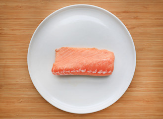 Raw piece of sliced salmon on white plate against wooden board background.