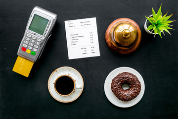 Electronic payments. Pay the bill by card concept. Bank card inserted in payment terminal near bill, service bell, coffee on black background top view