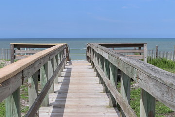 Fototapeta na wymiar Perspective photograph of wood boardwalk railing beach access horizon blue sky, turquoise ocean and green grass.