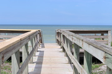 Obraz premium Perspective photograph of wood boardwalk railing beach access horizon blue sky, turquoise ocean and green grass.