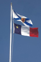 Chéticamp, Cape Breton, Nova Scotia, Canada: Flags of Nova Scotia (top) and Acadia (bottom), waving in the wind on flagpoles against a deep blue cloudless sky.