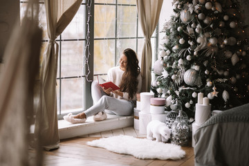 Nice dark-haired girl dressed in pants, sweater and warm slippers reads a book sitting on the...