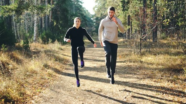 Happy young woman is doing sports with her handsome boyfriend jumping in park on path and laughing enjoying freedom, healthy activity and beautiful nature.