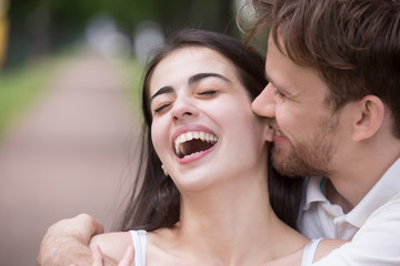 Loving man tickling and embracing laughing woman outdoors in park, tender boyfriend kissing or biting attractive girlfriend ear, telling funny news, story, family having fun together, close up