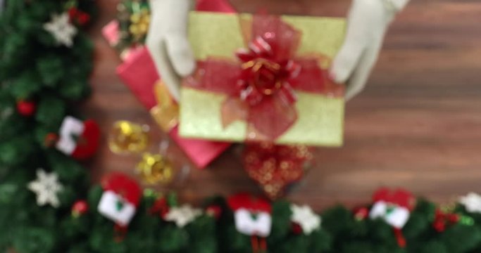 Close up scene VDO of blurred pine branches with beautiful ornament on wooden table, then has someone hands are holding gift box into frame and getting close to camera, then scene turn to be clear.