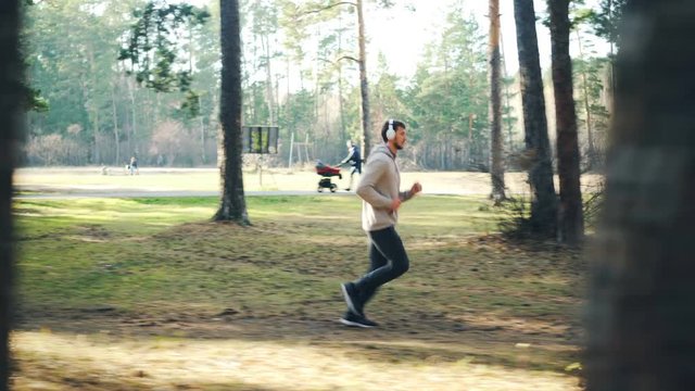 Dolly shot of bearded young man in tracksuit and sneakers jogging in park and listening to music through modern headphones. Sports and millennials concept.