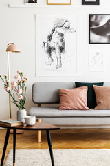 Flowers on wooden table in front of grey sofa with pillows in flat interior with posters. Real photo