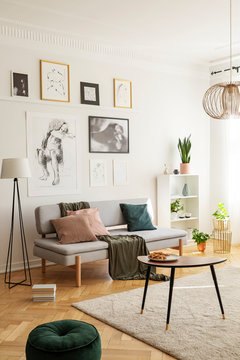 Posters above settee with pillows in bright living room interior with pouf next to table. Real photo