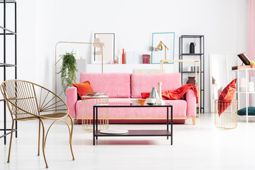 Gold armchair near table and pink sofa with orange blanket in white living room interior. Real photo
