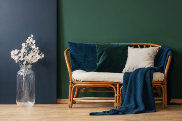 Blue blanket and pillows on rattan sofa next to white flowers in green apartment interior. Real photo