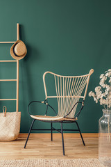 Flowers next to rattan armchair in green living room interior with hat on ladder above bag. Real...