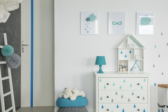 Posters above cabinet with blue lamp in white kid's room interior with plush toy on pouf. Real photo