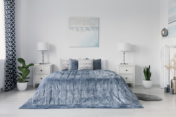 Blue bed between cabinet with lamps in white bedroom interior with plants and poster. Real photo