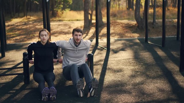 Young woman is exercising outdoors with her sporty boyfriend training arms using low parallel bars on sports-ground. Couple, sports and nature concept.