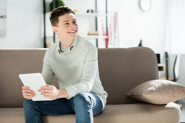 smiling teenager holding tablet on sofa at home and looking away