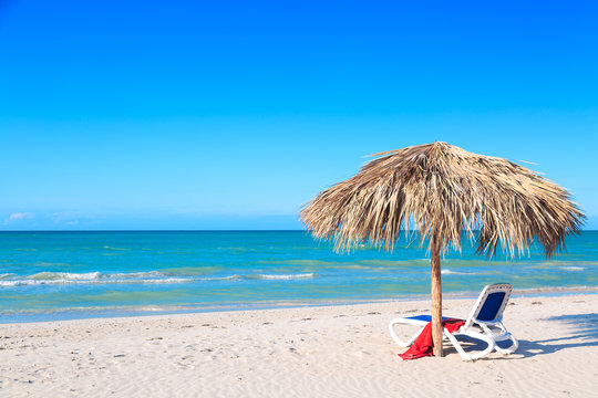 A sun lounger under an umbrella on the sandy beach by the sea and sky. Vacation background. Idyllic beach landscape.