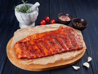 Marinated raw pork spare ribs on crumpled paper on a bamboo cutting board, with rosemary twigs, tomatoes, garlic cloves, pepper corns and coarse pink salt