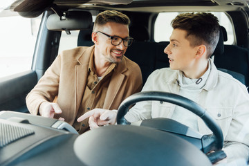 smiling handsome father teaching teen son driving car, they looking at each other and pointing on something