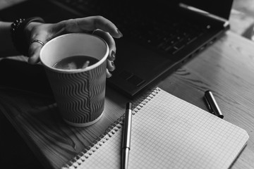woman hands and hot red cup of tea, coffee. Workplace, notebook with pen and laptop, the working process. coffee shop, space for text, morning work at the computer, study, black and white