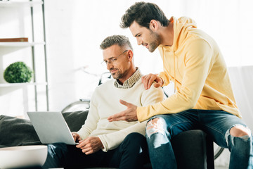 son gesturing and looking at mature father using laptop on sofa at home