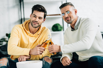 son and mature father clinking with bottles of beer on weekend at home and looking away