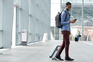 Full body side of young black man traveling with suitcase and cellphone at airport