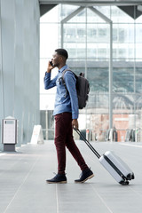 Full body side of young african american man traveling with suitcase and cellphone at airport